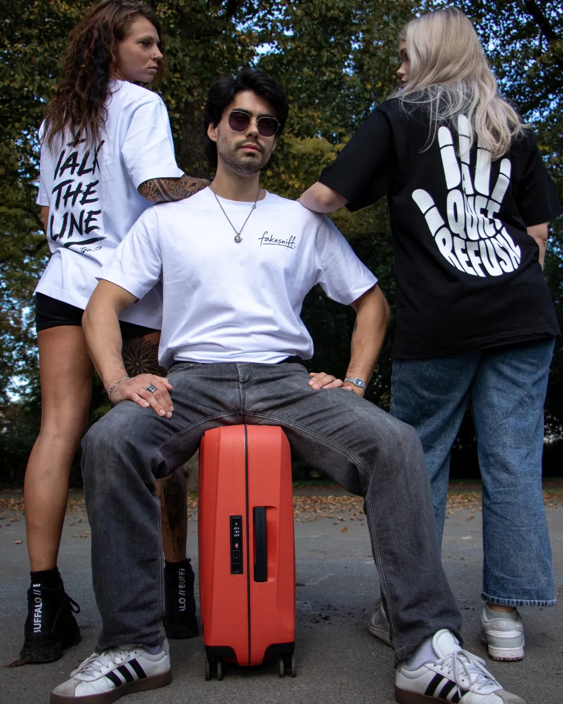 Three people on a basketball court. A man sits on a red suitcase, with two women beside him wearing I Walk The Line and A Quiet Refusal.