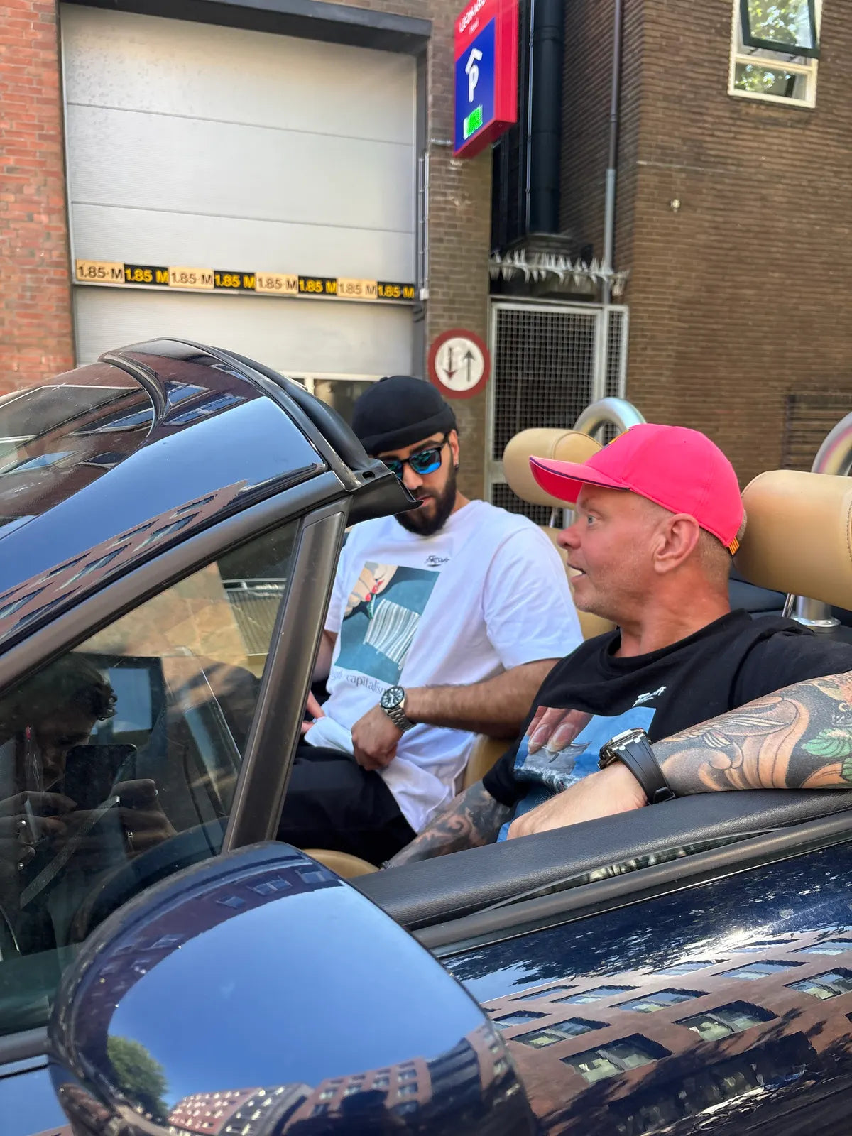 Two men in an open car in front of a car park.